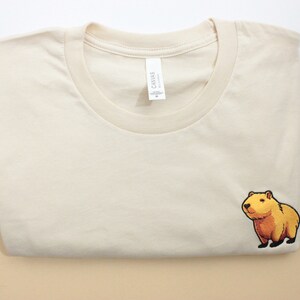 Short-sleeved soft cream T-shirt with a crew neckline, made from 100% combed and ring-spun cotton, features a small capybara embroidery on the left chest. The embroidery shows a standing small cartoon-style capybara capybara smiling, looking aside.