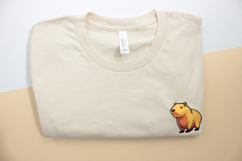 Short-sleeved soft cream T-shirt with a crew neckline, made from 100% combed and ring-spun cotton, features a small capybara embroidery on the left chest. The embroidery shows a standing small cartoon-style capybara capybara smiling, looking aside.