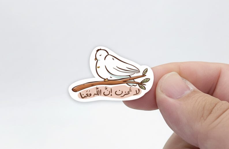 May include: A sticker featuring a cartoon white dove perched on a brown branch with green leaves. The sticker has Arabic text at the bottom. The dove is drawn with black outlines and yellow accents.