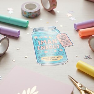 May include: A light blue sticker with the text "IMAN ENERGY" and a floral design, alongside colorful markers, floral patterned washi tape, a pink notebook, and gold scissors. The sticker reads "100% Halal Fuel."