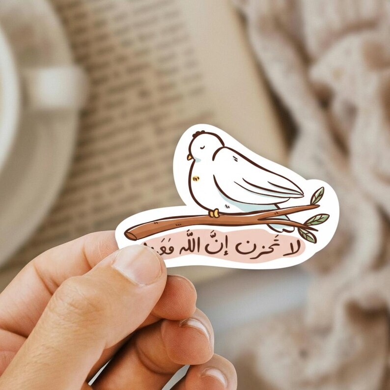 May include: A white sticker featuring a cartoon bird perched on a brown branch with green leaves. The sticker has Arabic text in a pink banner below the bird. The sticker is held by a hand.