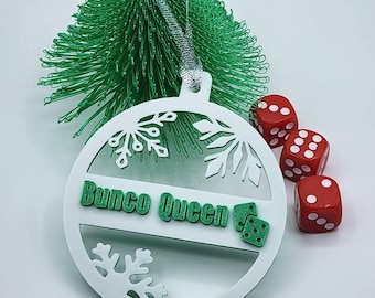 May include: White Christmas ornament with a green snowflake design and the text "Bunco Queen" in green. The ornament is hanging from a silver string and there are red dice in the background.