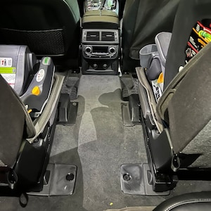 May include: A black car interior with two car seats in the back. The seats are folded down and the floor is carpeted. There is a small black box on the floor in the middle of the car.
