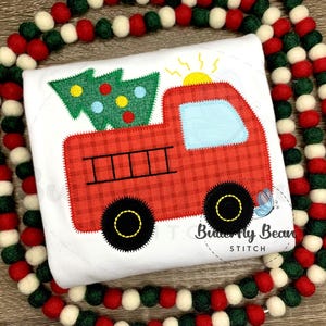 Può includere: Una maglietta bianca con un design di camion rosso a quadri che trasporta un albero di Natale verde. Il camion ha ruote nere, un finestrino blu e un sole giallo. Il design è incorniciato da una ghirlanda di palline di feltro rosse, bianche e verdi.
