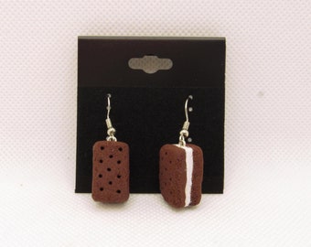 May include: A pair of dangle earrings featuring a chocolate cookie sandwich with white filling. The earrings are displayed on a black card with a silver hook.