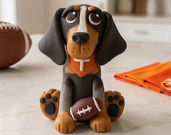 May include: A sculpted figurine of a black, brown, and white dog with large eyes, wearing an orange bandana with a white letter T. The dog is sitting and holding a small brown football with white laces between its front paws.