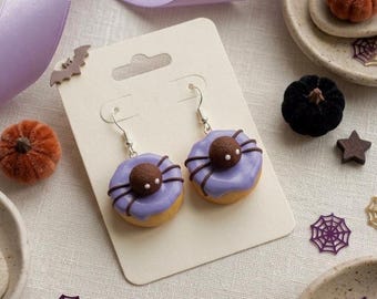 May include: A pair of dangle earrings featuring miniature donut charms with light purple icing and a small brown spider on top. The earrings are attached to a rectangular white display card with silver-toned metal hooks for pierced ears.