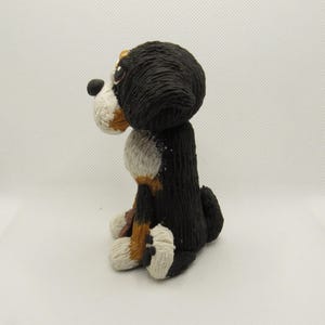 May include: A hand-painted ceramic figurine of a Bernese Mountain Dog sitting with its head turned to the side. The dog is black, white, and brown with a textured fur design.