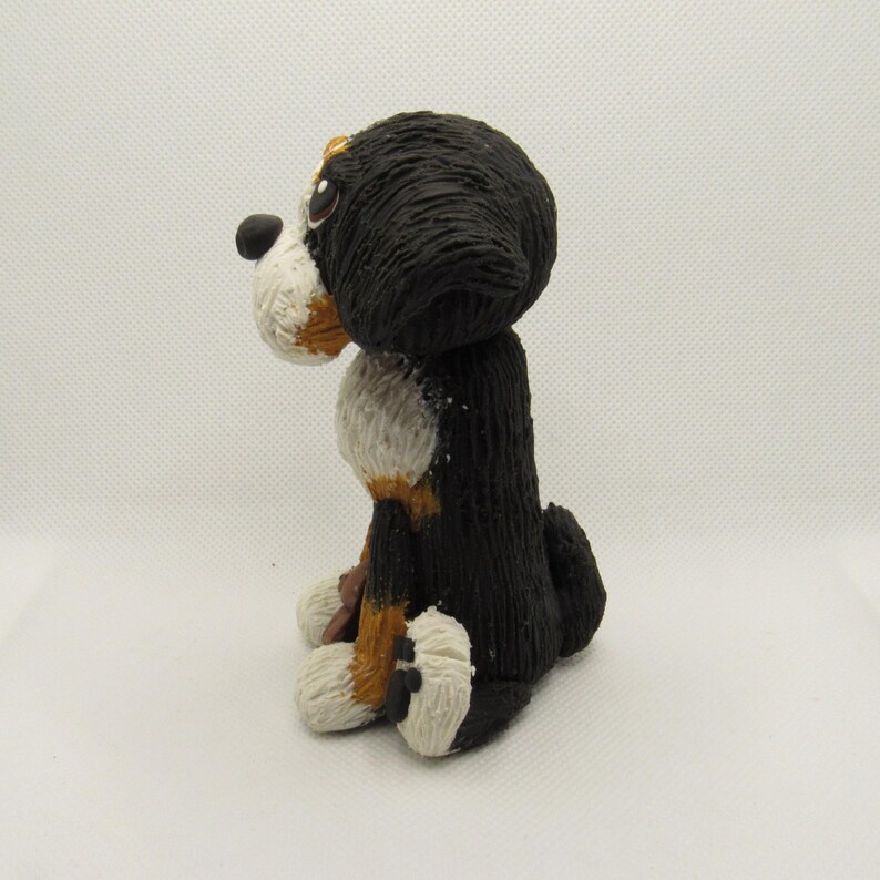 May include: A hand-painted ceramic figurine of a Bernese Mountain Dog sitting with its head turned to the side. The dog is black, white, and brown with a textured fur design.