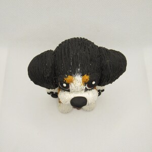 May include: A small, black, white, and brown ceramic figurine of a Bernese Mountain Dog puppy. The puppy is sitting with its head tilted down and its eyes closed.