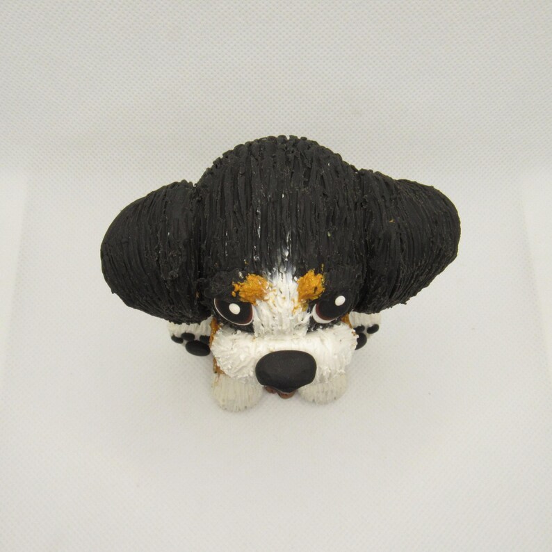 May include: A small, black, white, and brown ceramic figurine of a Bernese Mountain Dog puppy. The puppy is sitting with its head tilted down and its eyes closed.