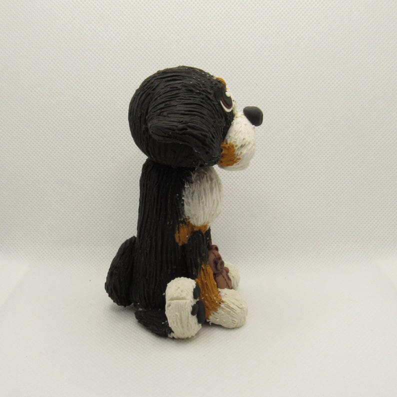 May include: A hand-painted ceramic figurine of a Bernese Mountain Dog sitting with its head turned to the side. The dog is black, white, and brown with a white patch on its chest.
