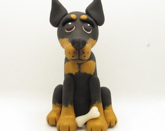 May include: A black and tan Doberman Pinscher figurine with a white bone in its paws. The figurine is sitting with its ears perked up and has a sad expression on its face.