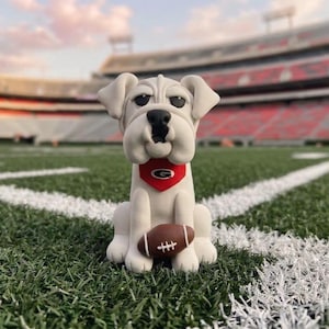 May include: A white bulldog figurine wearing a red bandana with a white "G" logo, holding a brown football. The figurine is sitting on a green football field with white yard lines, with a stadium in the background.