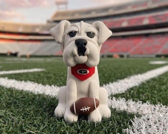 May include: A white bulldog figurine wearing a red bandana with a white "G" logo, holding a brown football. The figurine is sitting on a green football field with white yard lines, with a stadium in the background.