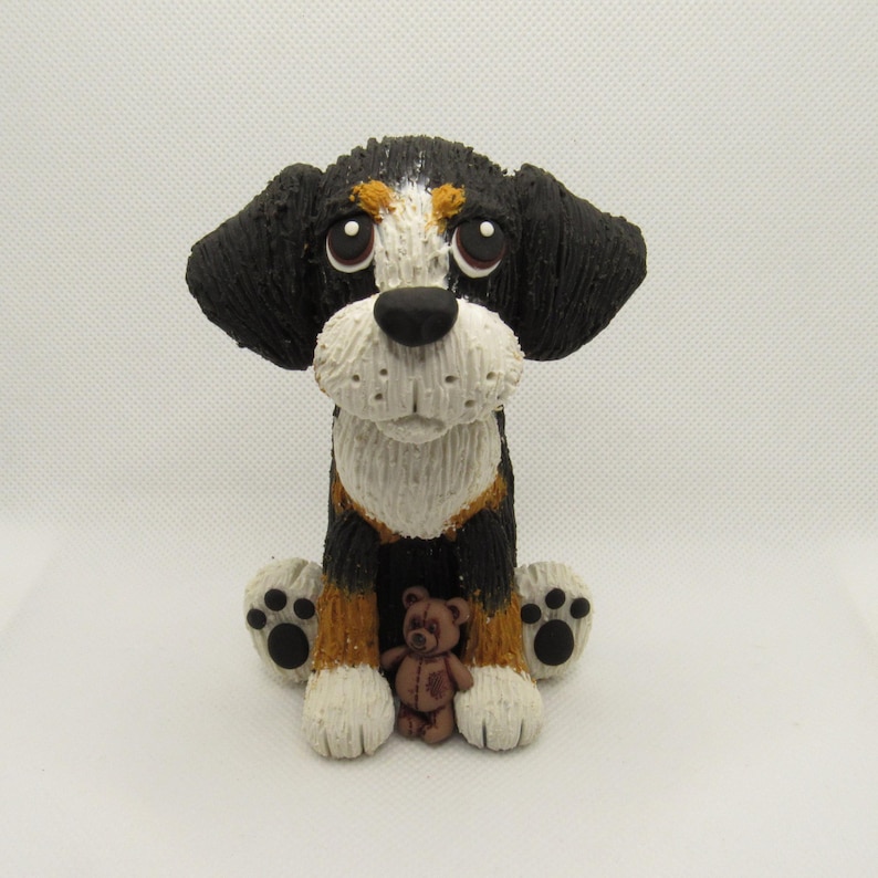 May include: A black, white, and brown ceramic figurine of a dog sitting with a brown teddy bear. The dog has big, brown eyes and a sad expression.