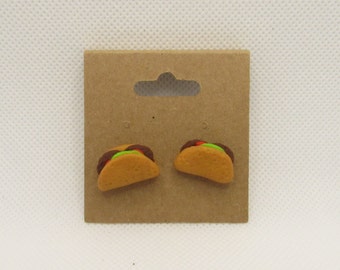 May include: A pair of taco-shaped earrings on a brown card. The earrings are made of polymer clay and are painted to look like tacos with brown shells, green lettuce, red tomatoes, and brown meat.