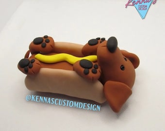 May include: A brown dachshund dog shaped like a hot dog with yellow mustard. The dog is lying on its back with its paws in the air. The hot dog is on a white background. The text "@KENNASCUSTOMDESIGN" is visible below the hot dog.