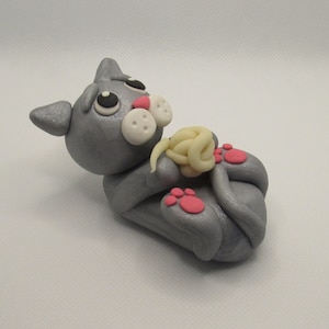 May include: A gray cat figurine with pink paws and a white nose, holding a ball of yarn in its paws.