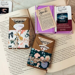 K&ouml;nnte beinhalten: Ein offenes Buch mit mehreren kleinen, farbenfrohen, buchf&ouml;rmigen Beh&auml;ltern und dekorativen Papierelementen. Die Beh&auml;lter sind in den Farbt&ouml;nen Gr&uuml;n, Orange, Lila und Rosa gehalten. Einige sind mit floralem Papier und Bindfaden umwickelt.