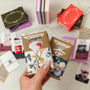 Puede incluir: Una colección de libros en miniatura en varios colores, incluyendo verde, rojo y morado. Dos libros están envueltos en papel floral y atados con hilo. El texto "Romantasy Chapter One" es visible en los envoltorios. Otros libros y efímeras están dispersos.