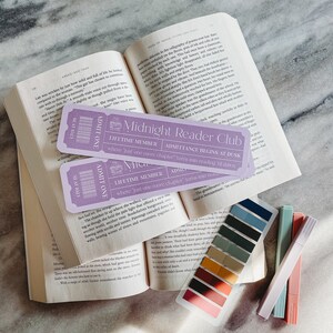 May include: Two purple bookmarks with the text "Midnight Reader Club" and "Lifetime Member" on a book with a colour palette of swatches. The bookmarks are for a book club.