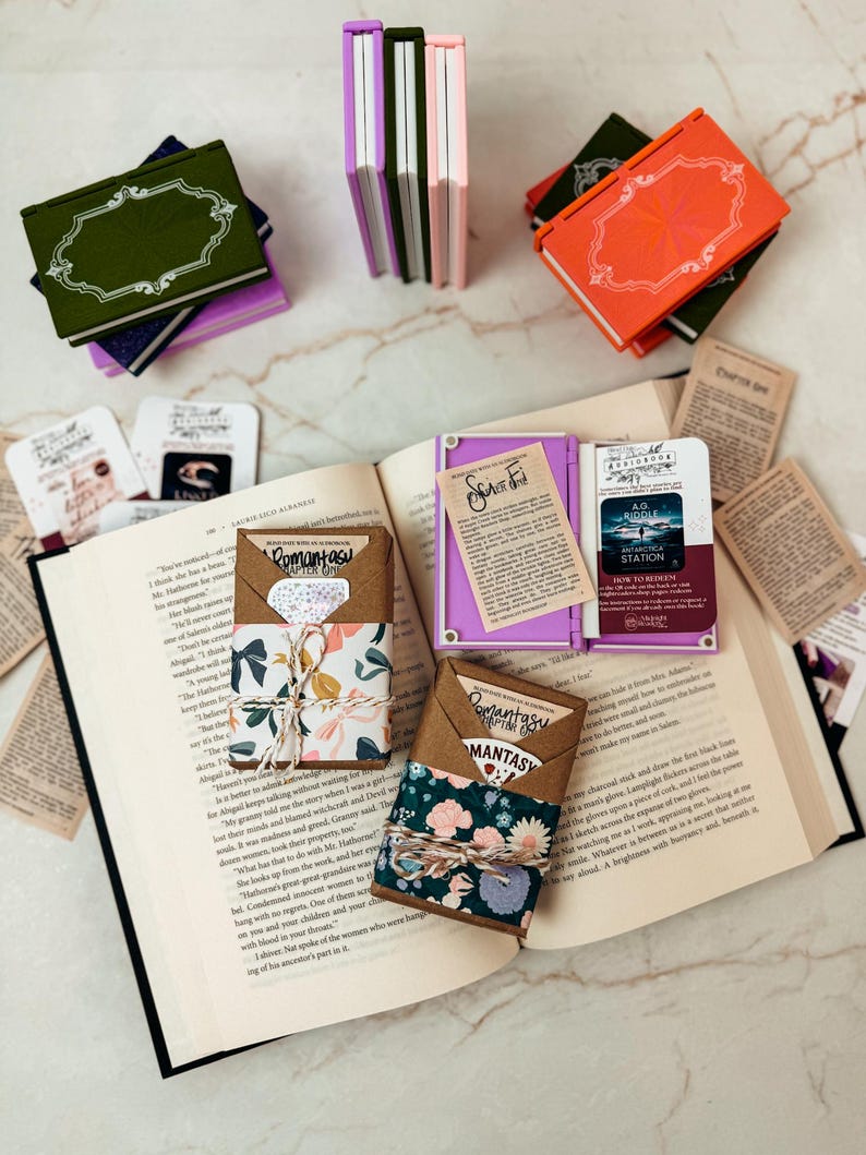 K&ouml;nnte beinhalten: Ein offenes Buch mit mehreren kleinen, farbenfrohen, buchf&ouml;rmigen Beh&auml;ltern und dekorativen Papierelementen. Die Beh&auml;lter sind in den Farbt&ouml;nen Gr&uuml;n, Orange, Lila und Rosa gehalten. Einige sind mit floralem Papier und Bindfaden umwickelt.