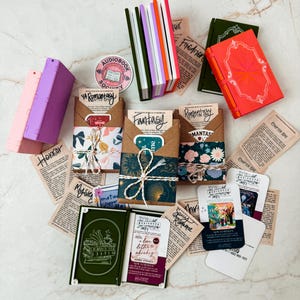 K&ouml;nnte beinhalten: Eine Auswahl an Miniaturb&uuml;chern und Artikeln zum Thema B&uuml;cher. Es gibt mehrere kleine, farbenfrohe B&uuml;cher, zusammen mit Papier-Ephemera und dekorativen Verpackungen zum Thema B&uuml;cher. Die Artikel sind auf einer hellen Oberfl&auml;che angeordnet.