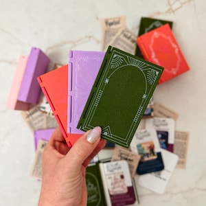 K&ouml;nnte beinhalten: Eine Hand h&auml;lt ein gr&uuml;nes, orangefarbenes und lila Miniaturbuch mit wei&szlig;en dekorativen Designs. Andere kleine B&uuml;cher in verschiedenen Farben und alte Papierschnipsel sind verstreut. Die B&uuml;cher haben ein Scharnierdesign.