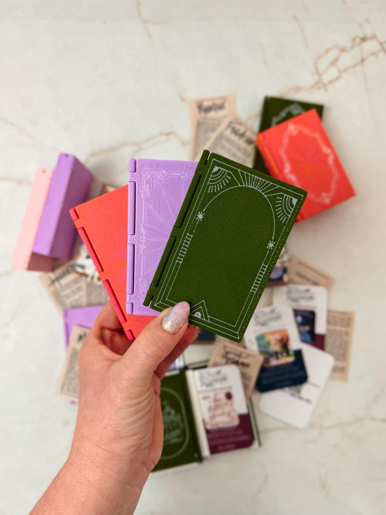 K&ouml;nnte beinhalten: Eine Hand h&auml;lt ein gr&uuml;nes, orangefarbenes und lila Miniaturbuch mit wei&szlig;en dekorativen Designs. Andere kleine B&uuml;cher in verschiedenen Farben und alte Papierschnipsel sind verstreut. Die B&uuml;cher haben ein Scharnierdesign.