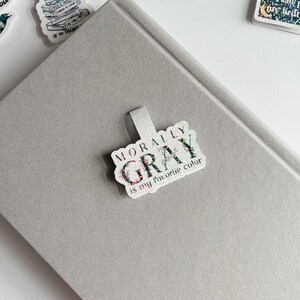 May include: A silver glitter bookmark with the text "Morally Gray is my favorite color" in black and white lettering.