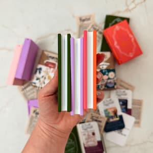 K&ouml;nnte beinhalten: Eine Sammlung farbenfroher Miniatur-Notizb&uuml;cher mit gr&uuml;nen, lila, rosa und orangefarbenen Buchr&uuml;cken. Die Notizb&uuml;cher werden in einer Hand gehalten, mit anderen kleinen B&uuml;chern und Papieren auf einer wei&szlig;en Marmoroberfl&auml;che verstreut.
