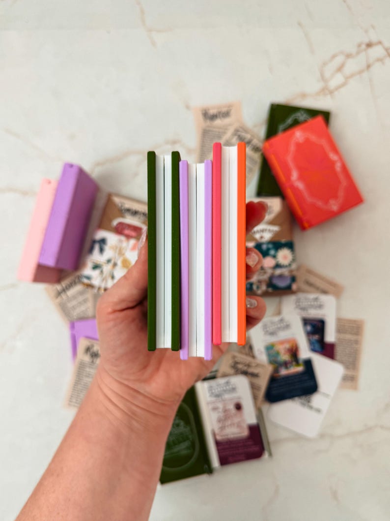 K&ouml;nnte beinhalten: Eine Sammlung farbenfroher Miniatur-Notizb&uuml;cher mit gr&uuml;nen, lila, rosa und orangefarbenen Buchr&uuml;cken. Die Notizb&uuml;cher werden in einer Hand gehalten, mit anderen kleinen B&uuml;chern und Papieren auf einer wei&szlig;en Marmoroberfl&auml;che verstreut.