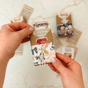 K&ouml;nnte beinhalten: Ein kleines, verpacktes P&auml;ckchen mit floralem und Schleifenmusterpapier, mit Bindfaden verschn&uuml;rt. Das P&auml;ckchen enth&auml;lt einen Buchauszug mit dem Text "YA Romantasy". Andere P&auml;ckchen und lose Papierschnipsel sind verstreut.