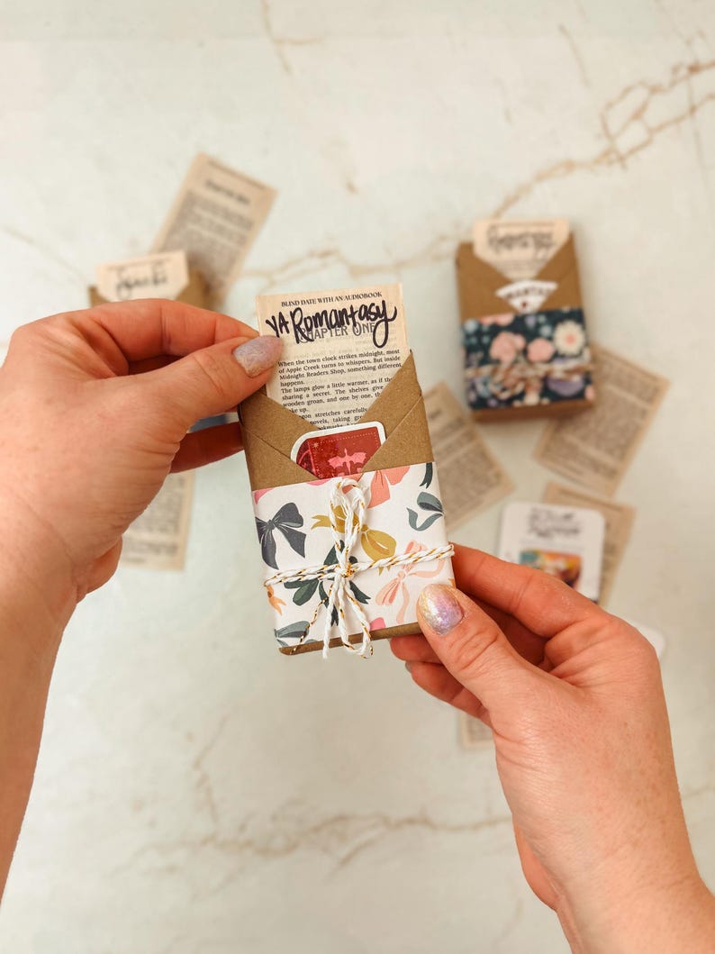 K&ouml;nnte beinhalten: Ein kleines, verpacktes P&auml;ckchen mit floralem und Schleifenmusterpapier, mit Bindfaden verschn&uuml;rt. Das P&auml;ckchen enth&auml;lt einen Buchauszug mit dem Text "YA Romantasy". Andere P&auml;ckchen und lose Papierschnipsel sind verstreut.