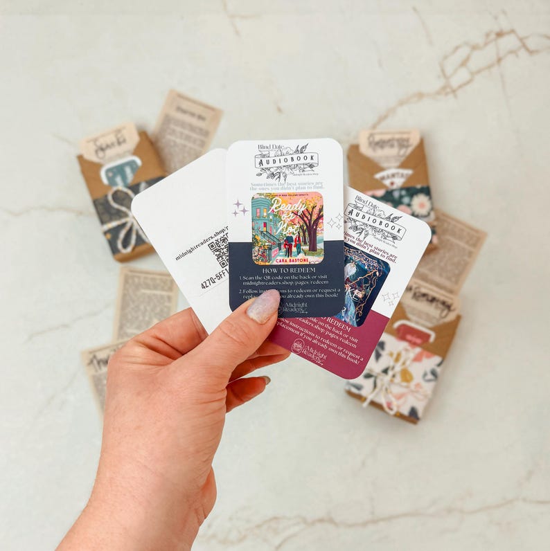 K&ouml;nnte beinhalten: Eine Hand h&auml;lt mehrere H&ouml;rbuchkarten mit einer farbenfrohen Illustration eines Hauses und eines Baumes. Die Karten tragen den Text "Blind Date with a Book" und "How to Redeem". Weitere Karten und kleine Pakete sind im Hintergrund zu sehen.