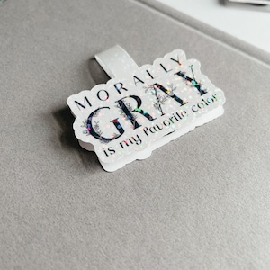May include: A white sticker with black text that reads "Morally Gray is my favorite color". The sticker has a glittery finish and floral accents.