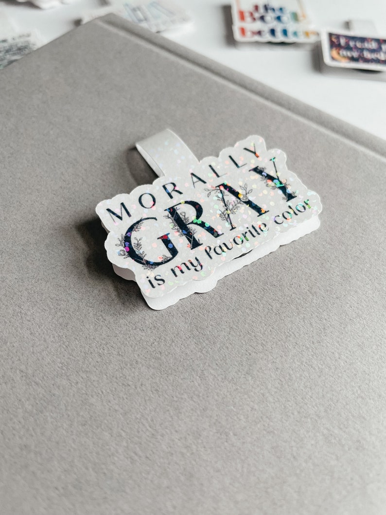 May include: A white sticker with black text that reads "Morally Gray is my favorite color". The sticker has a glittery finish and floral accents.