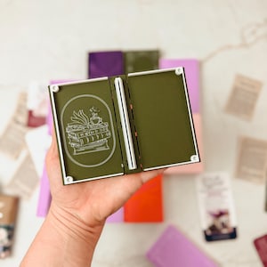 K&ouml;nnte beinhalten: Ein offenes, olivgr&uuml;nes, buchf&ouml;rmiges Portemonnaie mit wei&szlig;em Rand und einer wei&szlig;en Illustration von B&uuml;chern und einer Teetasse. Das Portemonnaie wird offen gehalten und zeigt einen wei&szlig;en Buchr&uuml;cken und ein leeres Innere. Andere farbige Geldb&ouml;rsen sind im Hintergrund.