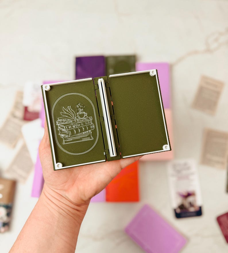 K&ouml;nnte beinhalten: Ein offenes, olivgr&uuml;nes, buchf&ouml;rmiges Portemonnaie mit wei&szlig;em Rand und einer wei&szlig;en Illustration von B&uuml;chern und einer Teetasse. Das Portemonnaie wird offen gehalten und zeigt einen wei&szlig;en Buchr&uuml;cken und ein leeres Innere. Andere farbige Geldb&ouml;rsen sind im Hintergrund.