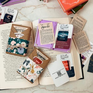 K&ouml;nnte beinhalten: Eine Ansammlung von B&uuml;chern und Zubeh&ouml;r liegt auf einer Marmoroberfl&auml;che. Offene B&uuml;cher, Lesezeichen und kleine, verpackte P&auml;ckchen mit floralem und gemustertem Papier sind zu sehen. Einige Artikel tragen Texte wie "Romantasy" und "Chapter One."