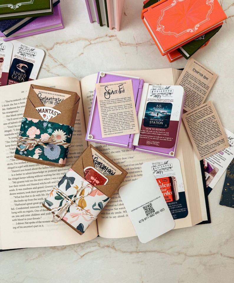 K&ouml;nnte beinhalten: Eine Ansammlung von B&uuml;chern und Zubeh&ouml;r liegt auf einer Marmoroberfl&auml;che. Offene B&uuml;cher, Lesezeichen und kleine, verpackte P&auml;ckchen mit floralem und gemustertem Papier sind zu sehen. Einige Artikel tragen Texte wie "Romantasy" und "Chapter One."