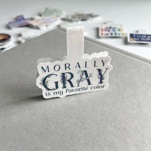May include: A white, glittery bookmark with the text "Morally Gray is my favorite color" in black lettering. The text is surrounded by a floral design.