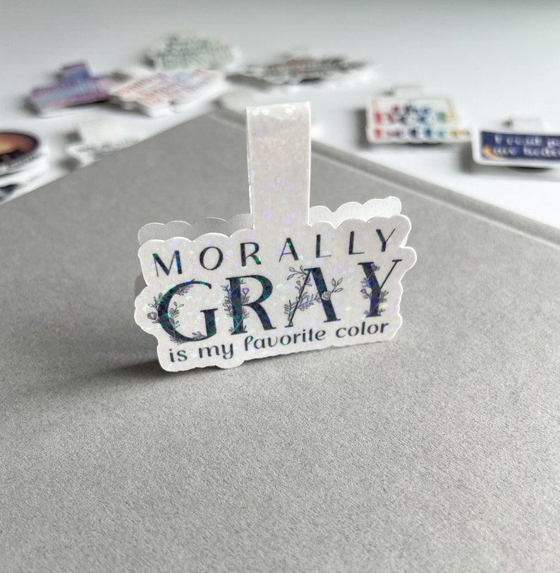May include: A white, glittery bookmark with the text "Morally Gray is my favorite color" in black lettering. The text is surrounded by a floral design.