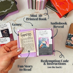K&ouml;nnte beinhalten: Eine Sammlung von Miniatur-3D-gedruckten B&uuml;chern in verschiedenen Farben, darunter Lila, Gr&uuml;n und Orange. Ein Buch ist ge&ouml;ffnet und zeigt eine gedruckte Geschichte und einen Einl&ouml;secode. Der Text im ge&ouml;ffneten Buch lautet "Romantasy" und "Sun StarMaker".