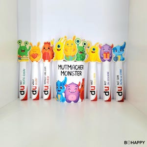 Könnte beinhalten: Eine weiße Tasse mit dem Text "MUTMACHER MONSTER" und bunten Monsterillustrationen. Die Tasse ist von Stifthaltern im Monster-Design umgeben. Jeder Stift hat ein anderes Monsterdesign und Text auf Deutsch.