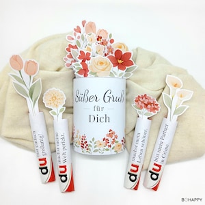May include: A white cylinder with floral designs and the text "Süßer Gruß für Dich" is surrounded by paper flowers and candy bars with German text. The flowers are in shades of peach, red, and white.