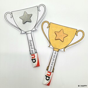 Könnte beinhalten: Zwei Pokal-Papierauschnitte, einer silberfarben und einer goldfarben, jeweils mit einem Stern und einem Griff aus einem Schokoriegel. Der silberne Pokal liest sich "bist großartig!" und der goldene Pokal liest sich "kannst stolz auf dich sein."