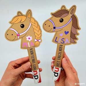 Może przedstawiać: Dwie ręcznie robione dekoracje w kształcie głowy konia z imionami "MIA" i "LISA". Głowy koni są brązowe z różowymi i fioletowymi akcentami. Na patyczkach widnieje napis "BIST EINGELADEN", a na dole mają czerwono-biały wzór.