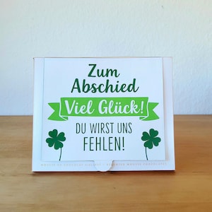 May include: A white box with a green and white label. The label has the text "Zum Abschied Viel Gl&uuml;ck! DU WIRST UNS FEHLEN!" and two green shamrock illustrations. The box is on a wooden surface.